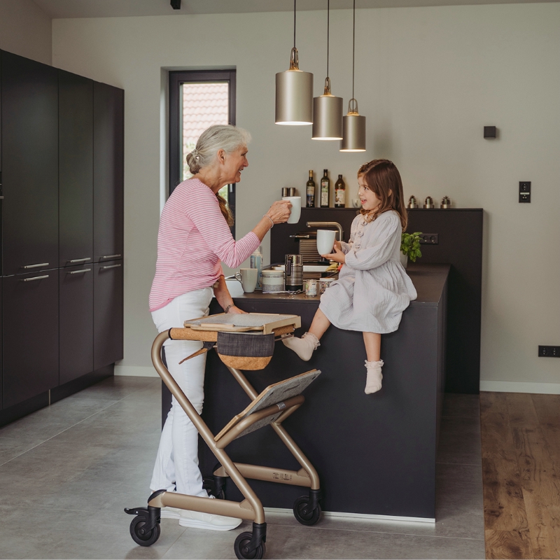 Femme âgée debout dans sa cuisine avec le rollator d'intérieur Saljol et son plateau, une petite fil